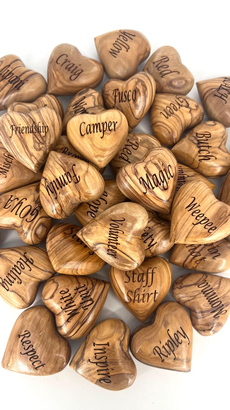 May include: A collection of smooth, heart-shaped wooden tokens crafted from olive wood, each featuring a unique grain pattern and a distinct word or name engraved in black text, including terms like Friendship, Camper, Magic, Family, and Respect.
