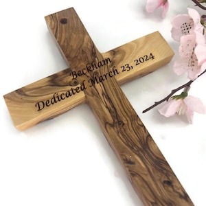 May include: Wooden cross with the inscription "Beckham Dedicated March 23, 2024." The cross is made of light brown wood with a visible grain pattern. Pink flowers are in the background.