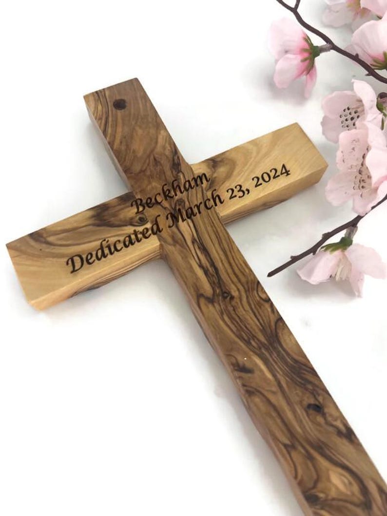 May include: Wooden cross with the inscription "Beckham Dedicated March 23, 2024." The cross is made of light brown wood with a visible grain pattern. Pink flowers are in the background.