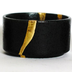 May include: A black ceramic bowl with gold accents. The bowl has a rough texture and is slightly wider at the top than the bottom.