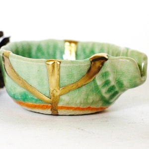 May include: A small, green ceramic bowl with gold accents and a textured surface. The bowl has a unique shape with a slight curve on one side. The bowl is sitting on a white surface.