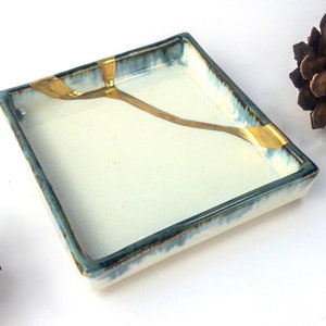 May include: A small, square ceramic dish with a white glaze and a blue rim. The dish has a gold stripe running across the bottom. The dish is sitting on a white surface.