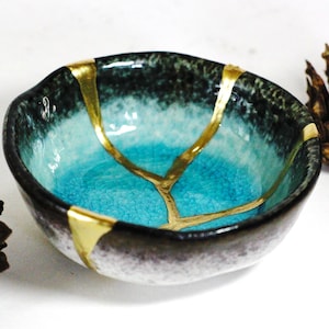 Puede incluir: Un pequeño cuenco redondo de cerámica con un interior azul turquesa y un exterior negro. El cuenco tiene detalles dorados que crean un efecto craquelado.