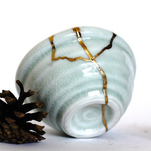 May include: A small, light blue ceramic bowl with gold cracks. The bowl is sitting on a white surface with two small pine cones on either side.