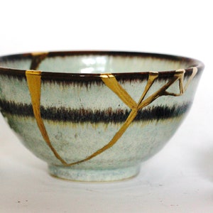May include: A small, light blue ceramic bowl with a brown rim and gold cracks. The bowl is sitting on a white surface with two pine cones on either side.