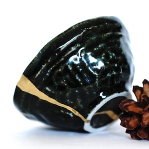 May include: A small, dark green ceramic bowl with gold and white accents. The bowl has a textured surface and is slightly chipped. A brown pine cone sits next to the bowl.
