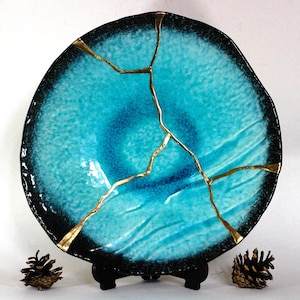 May include: A large, round, turquoise blue ceramic plate with gold crackle lines. The plate has a black rim and is on a black stand.