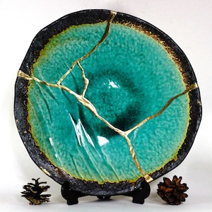 May include: A large, round, teal ceramic plate with a black rim and gold cracks. The plate is on a black stand and there are two pine cones on either side of the stand.