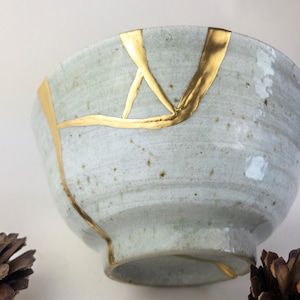 Könnte beinhalten: Eine weiße Keramikschüssel mit goldenen Rissen. Die Schüssel hat eine gesprenkelte Glasur und ist leicht abgebrochen.