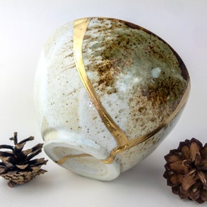 May include: A white ceramic bowl with a speckled brown and green glaze. The bowl has a gold rim and a gold line running down the side. The bowl is sitting on a white surface with two pine cones on either side.
