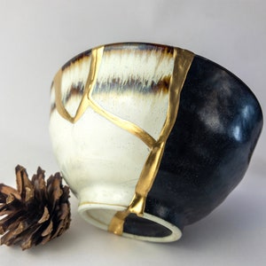May include: A white ceramic bowl with gold accents and a black glaze on one side. The bowl is slightly tilted and a small pine cone is in the foreground.