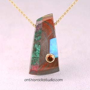 May include: A gold chain necklace with a pendant made of various stones, including red, green, blue, and brown. The pendant is set with a small, round, orange gemstone.