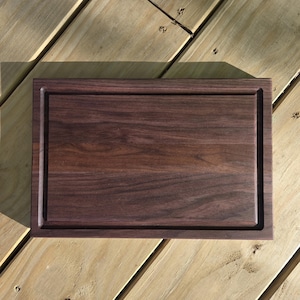 May include: A rectangular wooden cutting board with a dark brown finish. The board has a recessed border and a smooth surface. The wood grain is visible, adding texture. The board is designed for food preparation and serving.