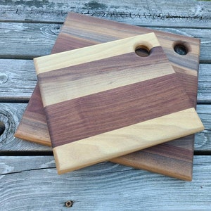 May include: Two wooden cutting boards, one on top of the other. The top board is smaller and has a light wood and dark wood striped pattern. The bottom board is larger and is a solid dark wood. Both boards have a hole in the top corner.