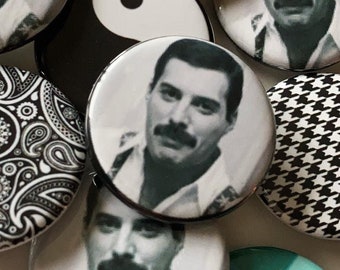 Könnte beinhalten: Eine Sammlung runder Ansteckbuttons mit verschiedenen Designs. Mehrere Buttons zeigen ein Schwarz-Weiß-Bild eines Mannes mit Schnurrbart. Andere Buttons haben ein Yin-Yang-Symbol, ein Paisley-Muster und ein Queen-Logo.