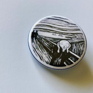 May include: A round button featuring a black and white print of Edvard Munch's famous artwork, "The Scream." The image depicts a figure with a wide-open mouth and hands on their face, set against a swirling landscape. The button has a white border with a thin blue line.
