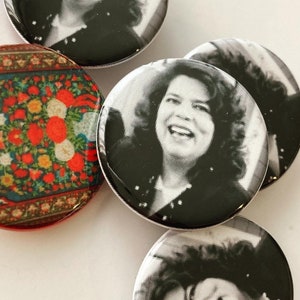 May include: A collection of round buttons. Four buttons feature a black and white image of a smiling person. One button has a colorful floral pattern with red, blue, and yellow hues. The buttons are arranged on a white surface.