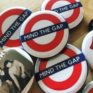 May include: A collection of round pinback buttons with various designs. Several feature the iconic London Underground symbol with the text "MIND THE GAP" on a blue banner. Other buttons display a band photo and the Rolling Stones' tongue logo.