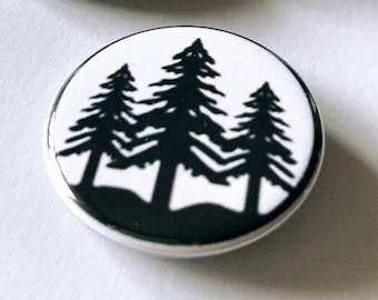 May include: A collection of four round, white buttons with a black silhouette of pine trees. The buttons have a black border and are arranged on a white surface. The design is simple and graphic.