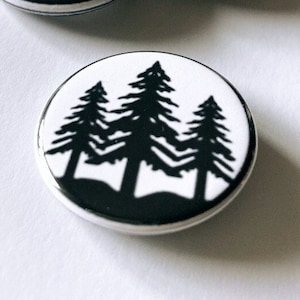 May include: A collection of four round, white buttons with a black silhouette of pine trees. The buttons have a black border and are arranged on a white surface. The design is simple and graphic.