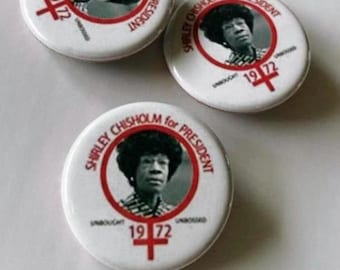 May include: Three white round buttons featuring a portrait of Shirley Chisholm, with the text "SHIRLEY CHISHOLM for PRESIDENT" in a red ring. The buttons also display "1972" and a red cross symbol. The buttons are on a white surface.