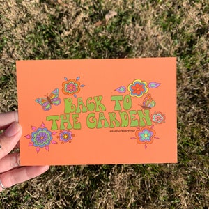 May include: An orange card with the text "Back to the Garden" in green, outlined in yellow. The text is in a retro font with flowers and butterflies in pink, blue, green, and yellow.