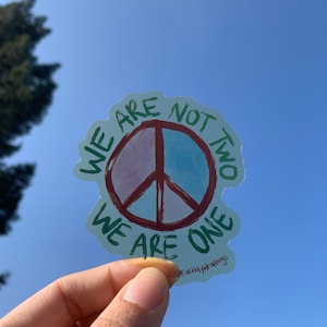 May include: A sticker with a peace sign design. The peace sign is divided into two sections, one pink and one blue, with a brown outline. The sticker has the text "WE ARE NOT TWO WE ARE ONE" in green. The sticker is held up against a blue sky.