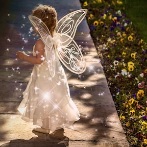 May include: A young girl wearing a white fairy costume with sparkling wings. She is walking on a stone path with a flower garden in the background.
