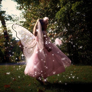 May include: A young girl dressed as a fairy with pink tulle wings and a pink dress, holding a wand with a pink flower on the end. She is standing in a grassy area with trees in the background.