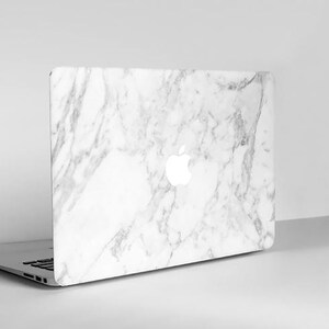 Puede incluir: Un ordenador port&aacute;til con una cubierta con dise&ntilde;o de m&aacute;rmol blanco. El logotipo de Apple es visible en la parte posterior. El port&aacute;til est&aacute; abierto e inclinado ligeramente hacia la izquierda, mostrando el teclado.