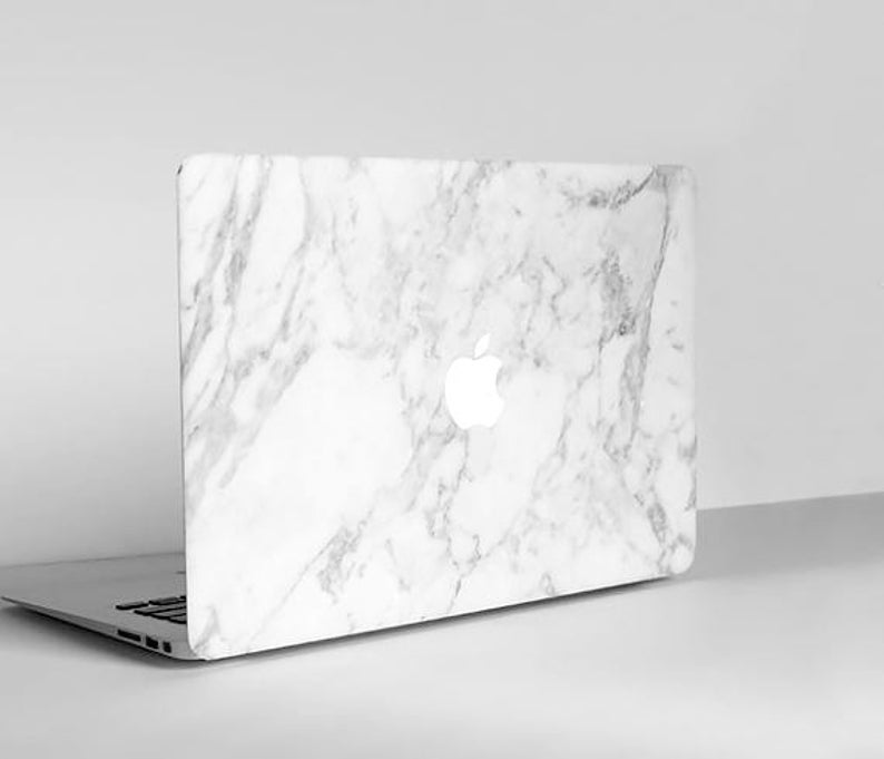 Puede incluir: Un ordenador port&aacute;til con una cubierta con dise&ntilde;o de m&aacute;rmol blanco. El logotipo de Apple es visible en la parte posterior. El port&aacute;til est&aacute; abierto e inclinado ligeramente hacia la izquierda, mostrando el teclado.