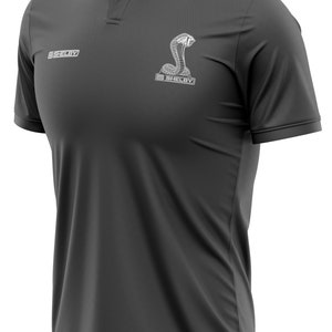 Puede incluir: Una camiseta polo gris oscuro con un logotipo Shelby bordado en blanco y un dise&ntilde;o de serpiente. El logotipo dice "Shelby" y la serpiente es una cobra.