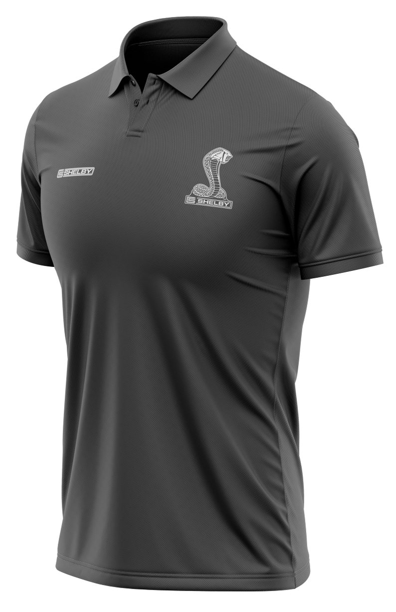 Puede incluir: Una camiseta polo gris oscuro con un logotipo Shelby bordado en blanco y un dise&ntilde;o de serpiente. El logotipo dice "Shelby" y la serpiente es una cobra.