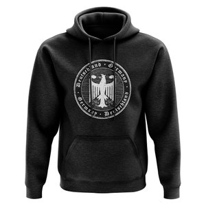 Może przedstawiać: Ciemnoszara bluza z kapturem z biało-czarnym niemieckim emblematem orła. Emblemat jest okrągły i zawiera słowa "Deutschland" i "Germany". Bluza z kapturem ma kaptur i przednią kieszeń.