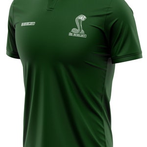 Puede incluir: Una camiseta polo verde oscuro con un logotipo blanco bordado de Shelby Cobra en el pecho. El logotipo presenta una serpiente con la capucha levantada y la palabra "Shelby" debajo.