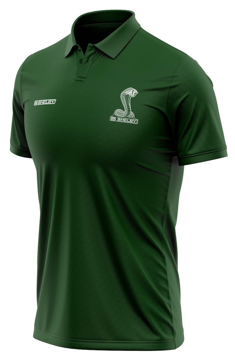 Puede incluir: Una camiseta polo verde oscuro con un logotipo blanco bordado de Shelby Cobra en el pecho. El logotipo presenta una serpiente con la capucha levantada y la palabra "Shelby" debajo.