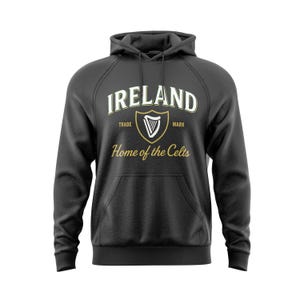 Puede incluir: Sudadera con capucha gris oscuro con la palabra "IRELAND" en blanco arqueada en la parte superior. Debajo, un escudo dorado con un arpa y el texto "Home of the Celts". La sudadera tiene un bolsillo delantero y una capucha.