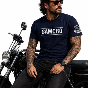 Peut inclure: T-shirt bleu marine avec le texte "SAMCRO" dans un rectangle blanc. Le t-shirt présente également un motif sur la manche. La personne sur l'image porte des lunettes de soleil noires et un jean noir, et est assise sur une moto noire.