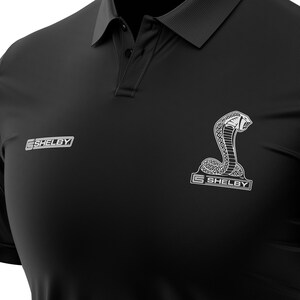 Puede incluir: Polo negro con un logotipo Shelby blanco y un gr&aacute;fico de serpiente plateado. El logotipo dice "Shelby" con un s&iacute;mbolo de marca registrada.