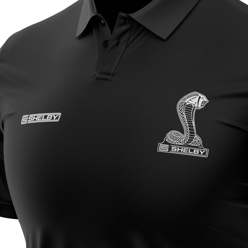 Puede incluir: Polo negro con un logotipo Shelby blanco y un gr&aacute;fico de serpiente plateado. El logotipo dice "Shelby" con un s&iacute;mbolo de marca registrada.