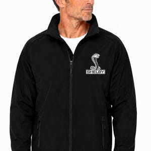 May include: A black zip-up jacket featuring a high collar and long sleeves. The left chest displays a white embroidered logo of a cobra snake above a rectangular box containing the text SHELBY. The jacket includes two side pockets with zippers.