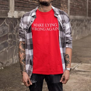 May include: A red t-shirt with the text "MAKE LYING WRONG AGAIN" printed in white. The person wearing the shirt is wearing a plaid shirt over it.