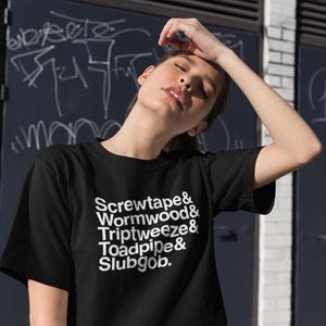 May include: Black t-shirt with white text: "Screwtape & Wormwood & Triptweeze & Toadpipe & Slubgob." The shirt is short-sleeved and made of a soft material. The background features a dark wall with graffiti and a white brick wall.