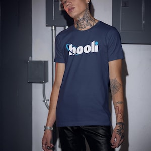 May include: Navy blue t-shirt with the word "hooli" in white and blue. The shirt is paired with black leather pants. The image is taken indoors with industrial-style electrical boxes in the background.