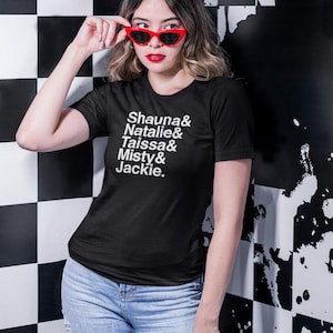 以下が含まれることがあります： 黒いTシャツを着た女性。Tシャツには「Shauna & Natalie & Taissa & Misty & Jackie.」の文字が書かれている。赤いキャットアイのサングラスと青いジーンズを着用している。