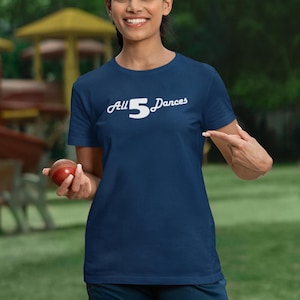 Pode incluir: Uma mulher vestindo uma camiseta azul marinho com o texto "All 5 Dances" em branco. Ela está segurando uma bola de críquete vermelha e usando protetores de críquete brancos.