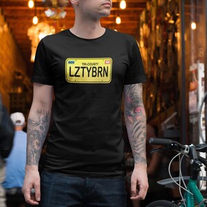 Op de afbeelding: Een zwart T-shirt met een gele kentekenplaat-afbeelding waarop "TRI-COUNTY LZTYBRN NOV" staat.