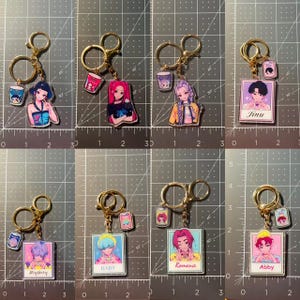 May include: A collection of eight anime-style keychains. Each keychain features a gold-colored ring and clasp, with a small charm of a character and a drink. The characters have different hair colors and outfits. Some keychains have names like "Jinu", "Mystery", "Baby", "Romance", and "Abby".