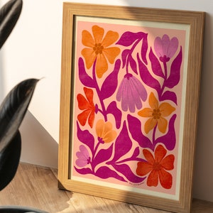 May include: A framed print with a pink background featuring a floral design with orange, red, and purple flowers. The flowers are stylized and have a retro feel. The print is framed in a light wood frame.