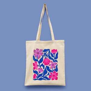 May include: A beige tote bag with a floral design. The design features pink and purple flowers with blue leaves. The tote bag has long beige handles and is made of a canvas-like material. The background is a solid light blue.